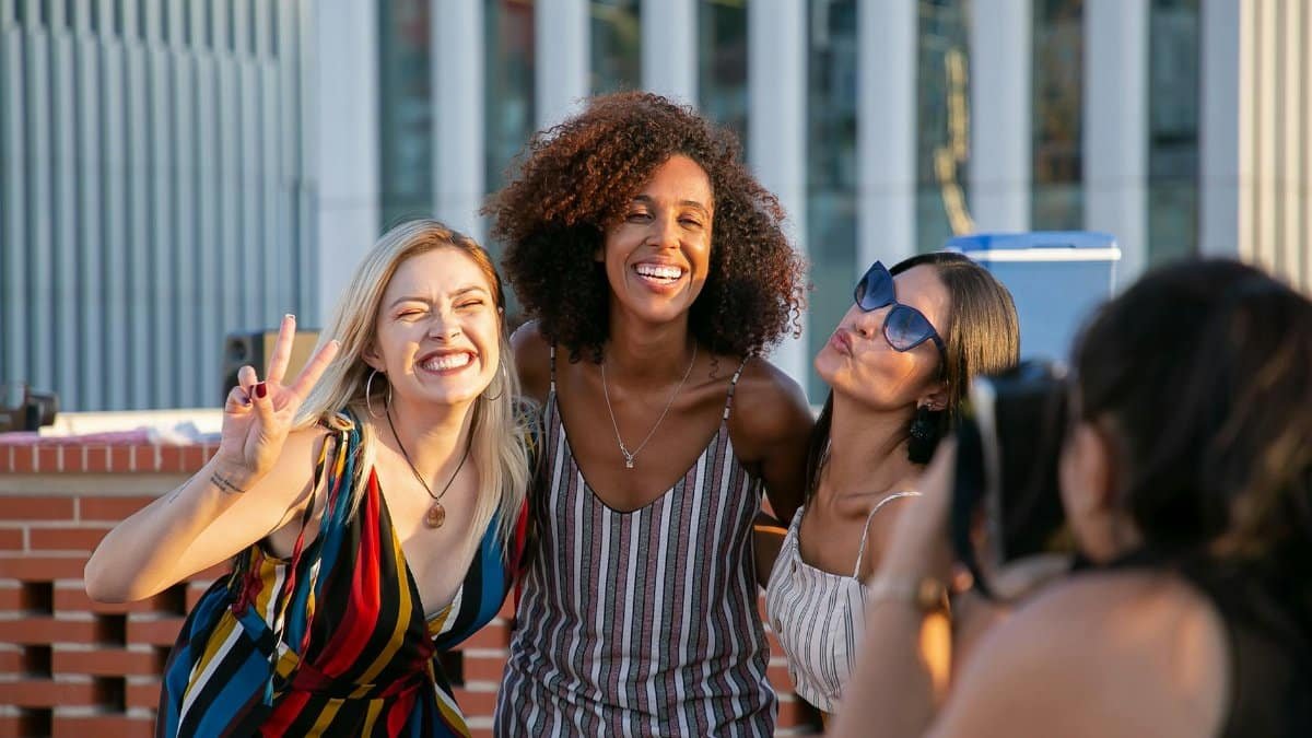Unrecognizable female photographer taking picture of cheerful young multiracial female friends smiling and showing V sign during party on modern building rooftop