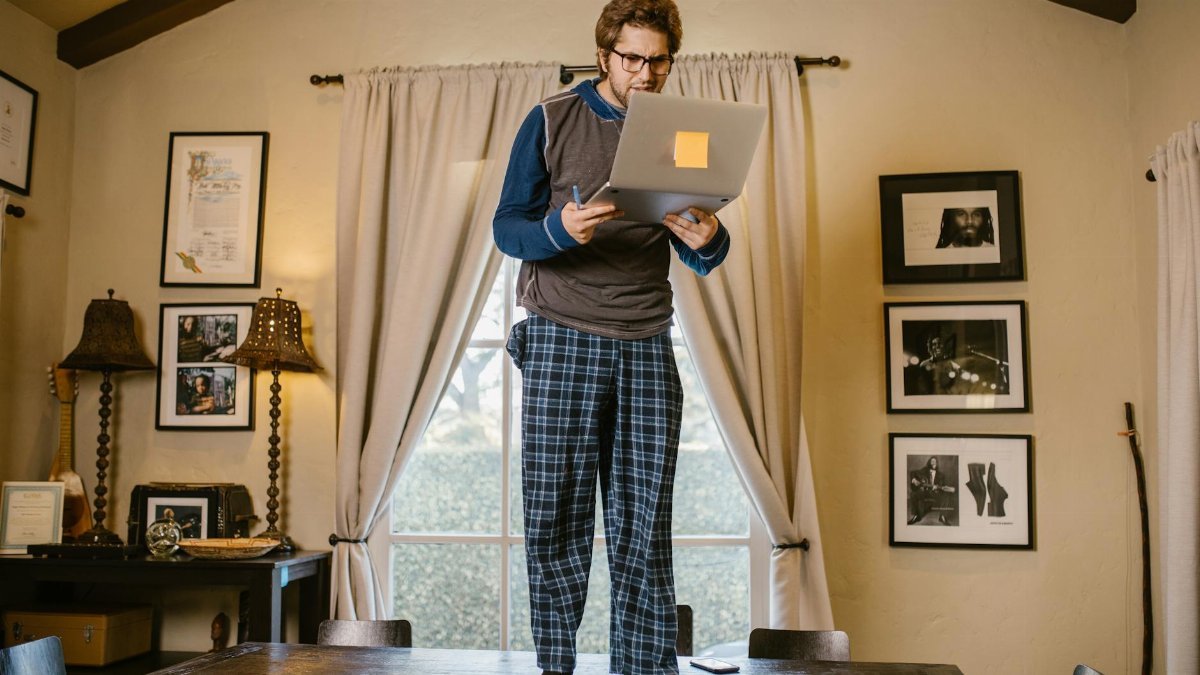 Man in pajamas standing on table with a laptop indoors, focused on remote work or study.