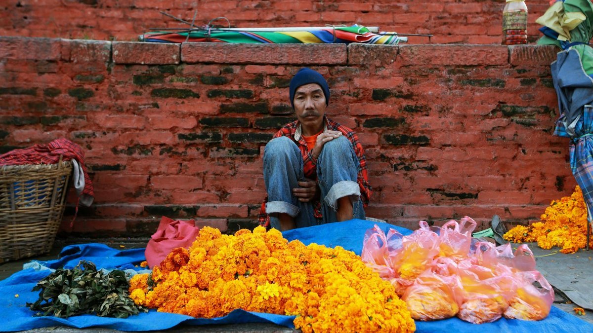 A street vendor sits by vibrant marigolds on display against a brick backdrop. Perfect capture of traditional market scene.