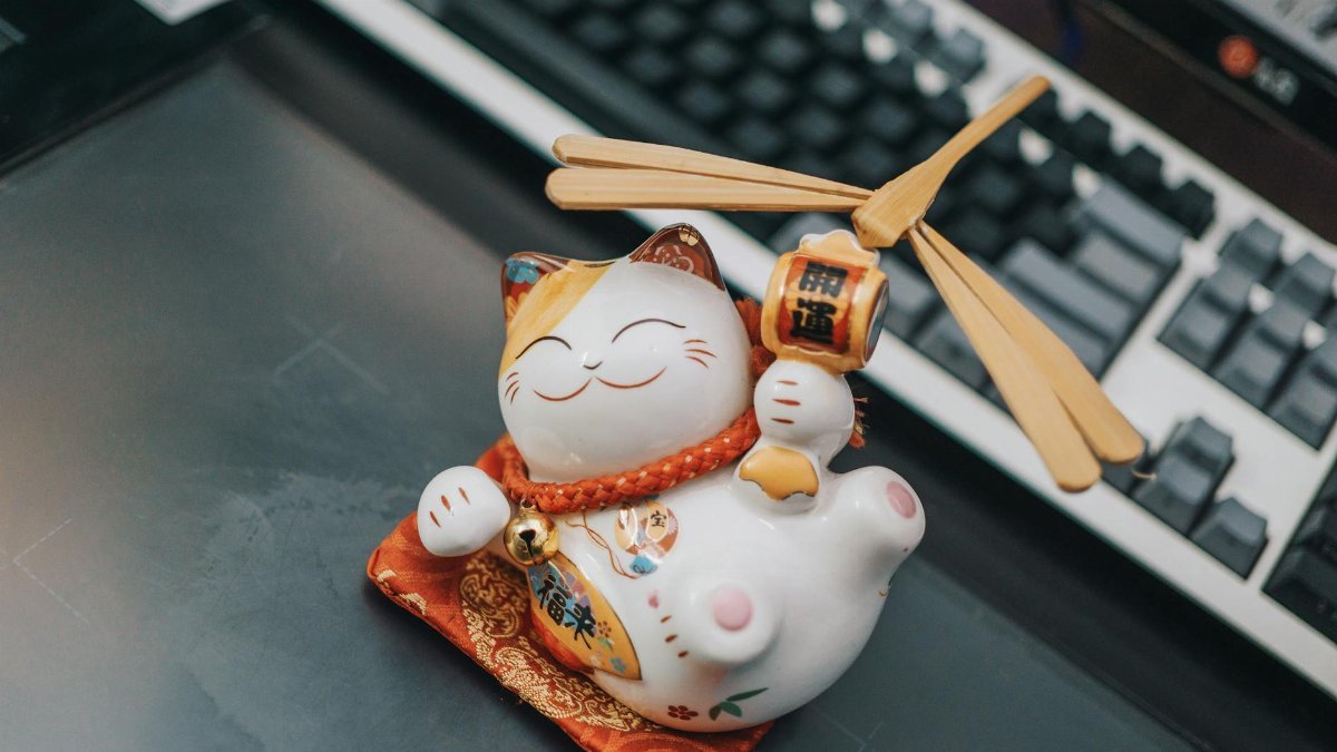 A ceramic Maneki Neko figurine sitting on a desk beside a keyboard, symbolizing luck and prosperity.