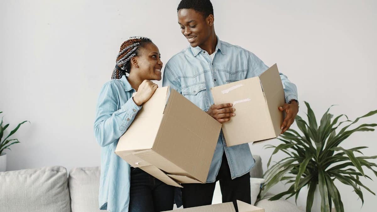 A smiling couple carrying cardboard boxes while moving indoors, capturing a joyful moment.
