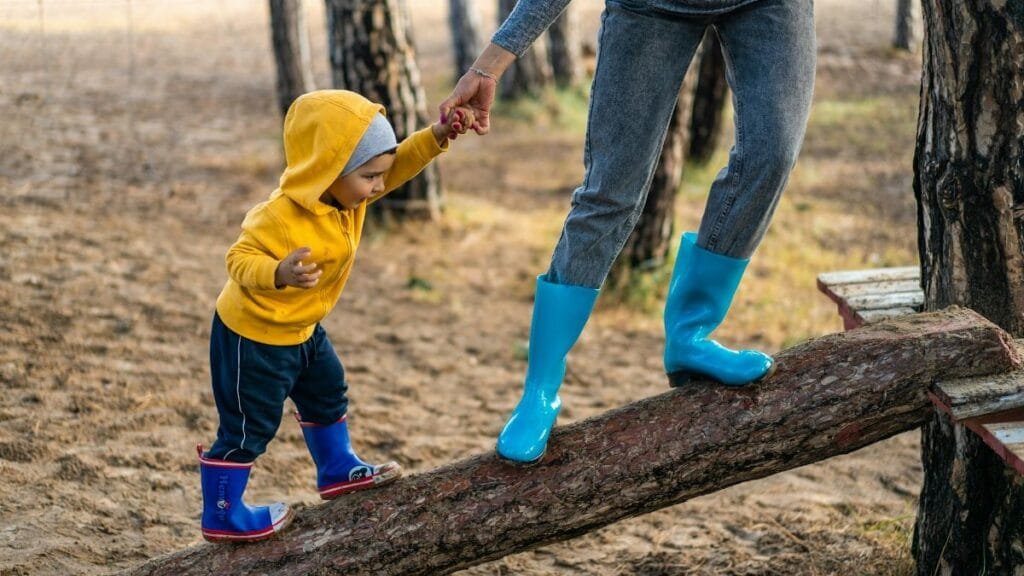 A woman helps her toddler walk on a log in a park, showcasing child support and family bonding.