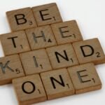 Close-up of wooden letter tiles forming the message 'Be the kind one' on a white surface.