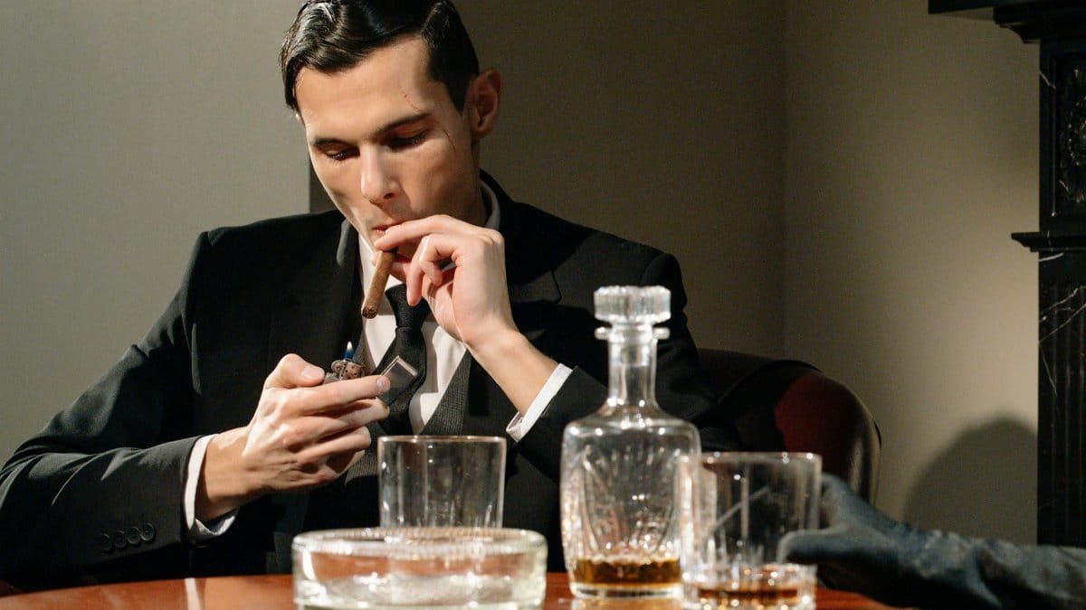 A sophisticated man in a suit smokes a cigar while enjoying a drink, exuding luxury and vintage style.