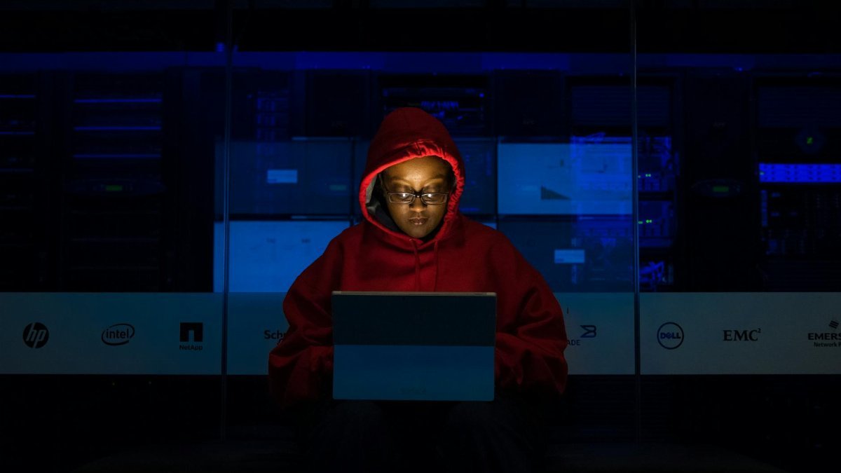 Focused woman in a red hoodie using a laptop in a dimly lit room with technology brands displayed.