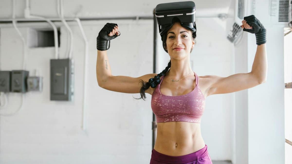 Woman flexes muscles with VR headset and gloves in gym, showcasing fitness technology.