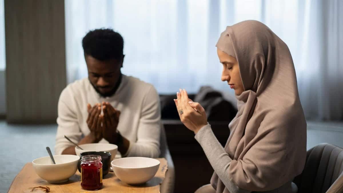 A couple sits indoors with hands raised in prayer before a meal, symbolizing spirituality.
