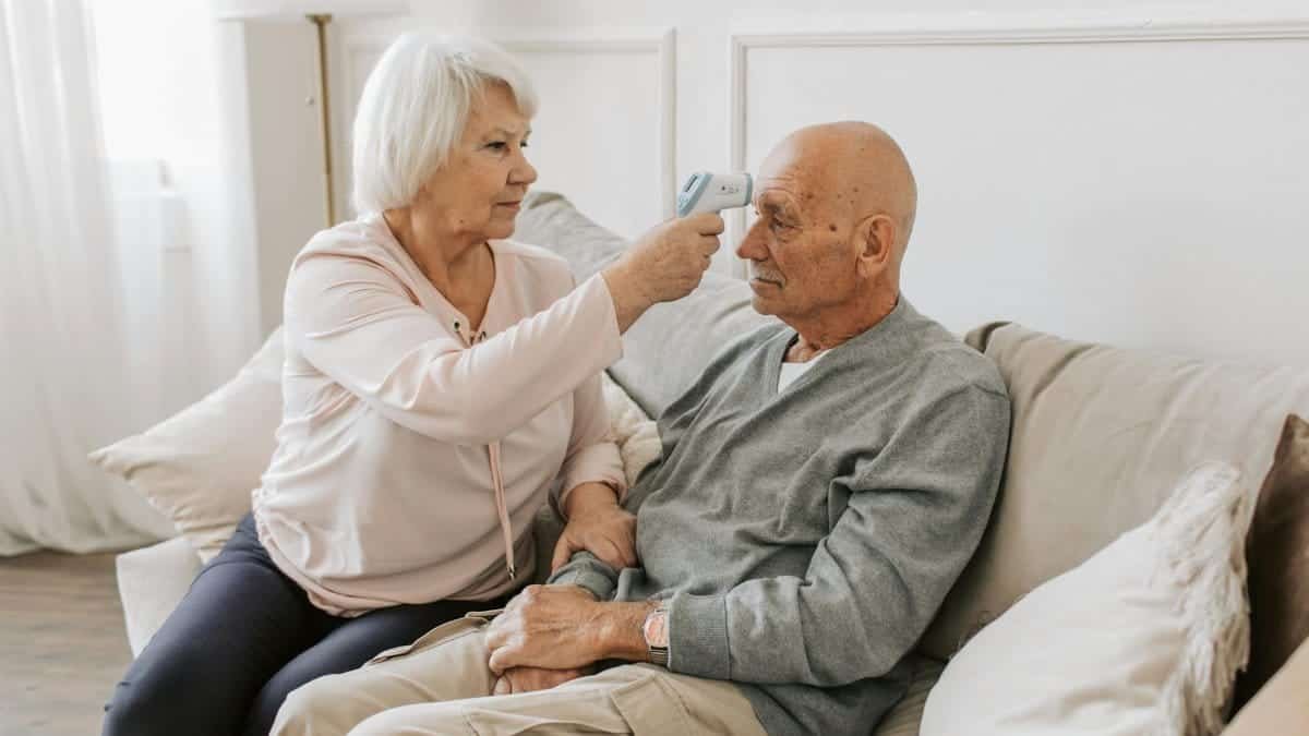 Senior woman checking her partner's temperature on a sofa, promoting health care.