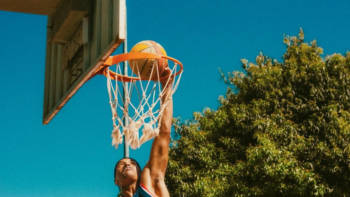 Young athlete performing a basketball dunk outdoors under a clear blue sky.