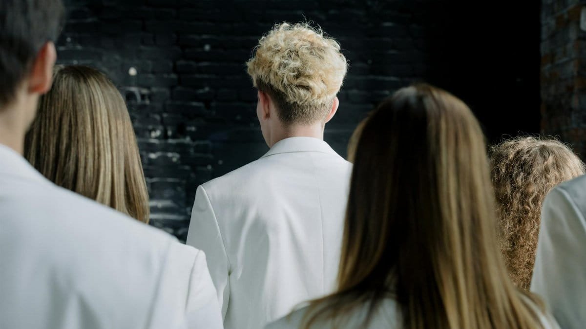 A group of people wearing white outfits with their backs turned, facing a dark wall.