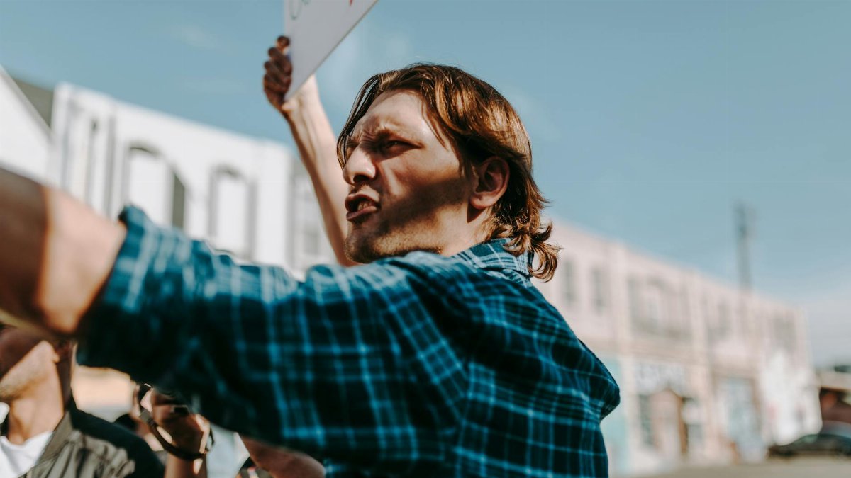 Man in a plaid shirt passionately speaking and holding a sign during an outdoor protest.
