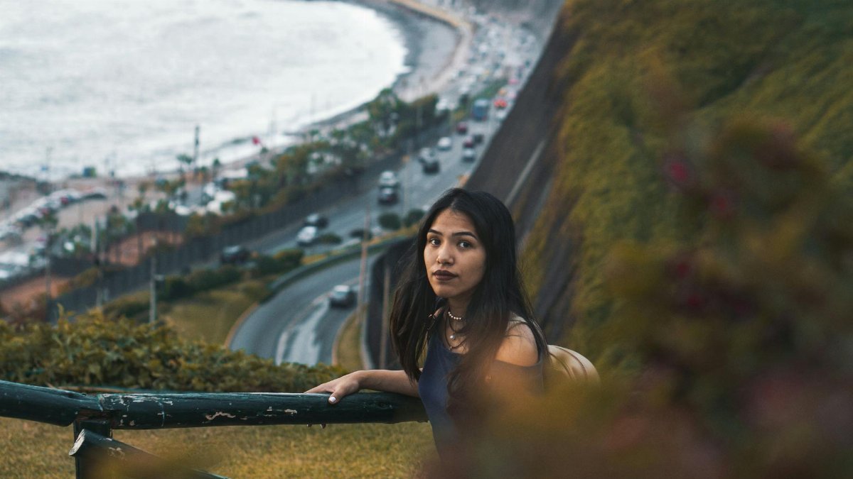 A woman leans on a railing with a scenic coastal highway backdrop.