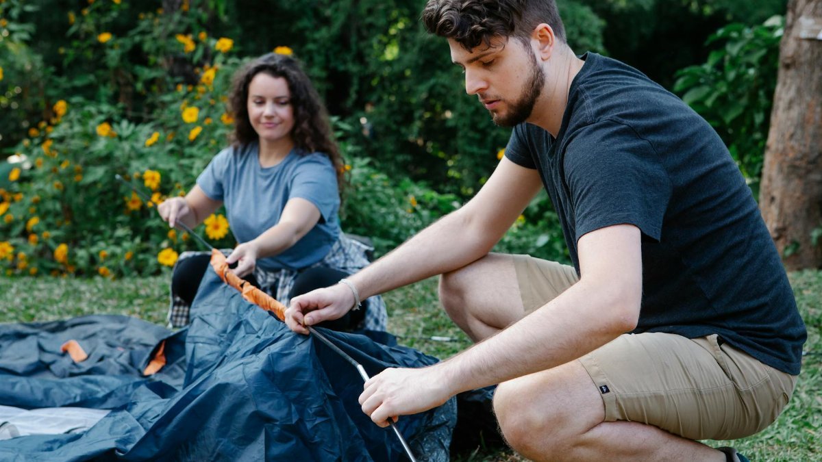 Concentrated couple of travelers in casual clothes squatting down and pitching tent in green forest during hike in daytime