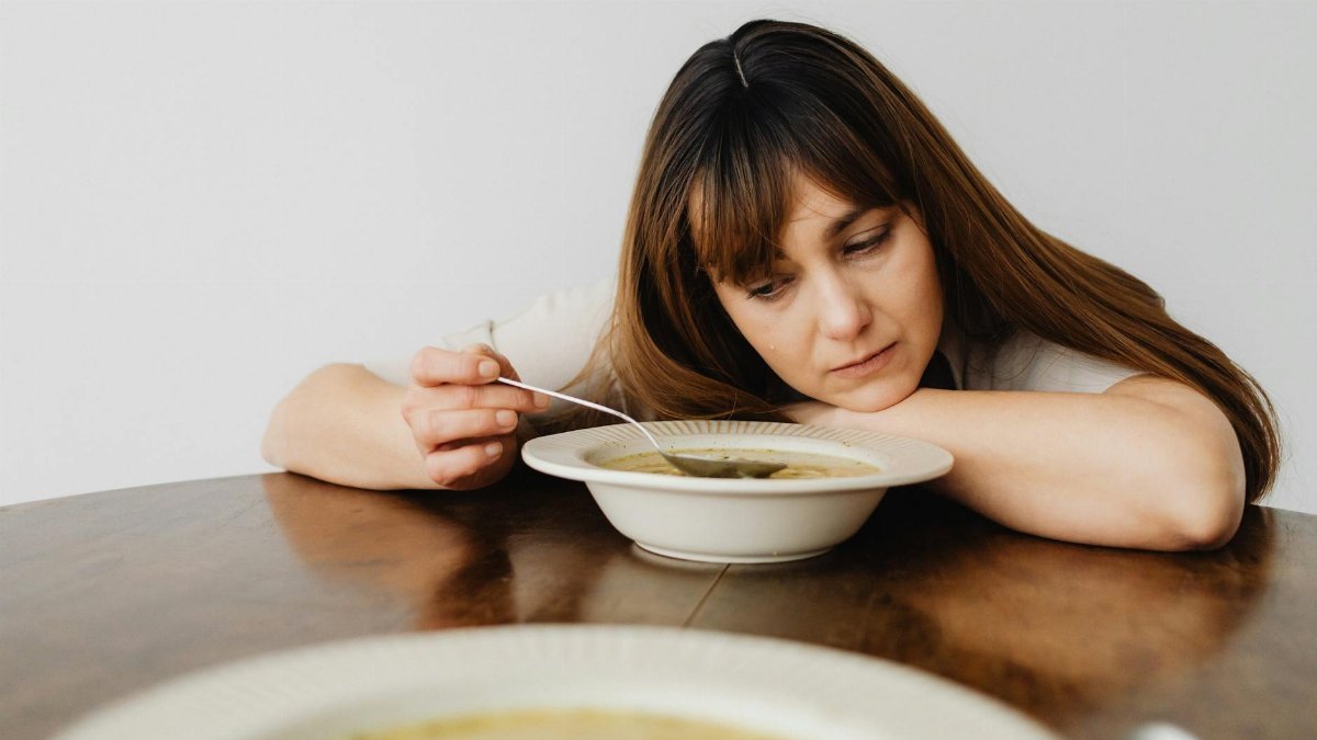 Woman with long hair looking bored while eating soup at a wooden table.
