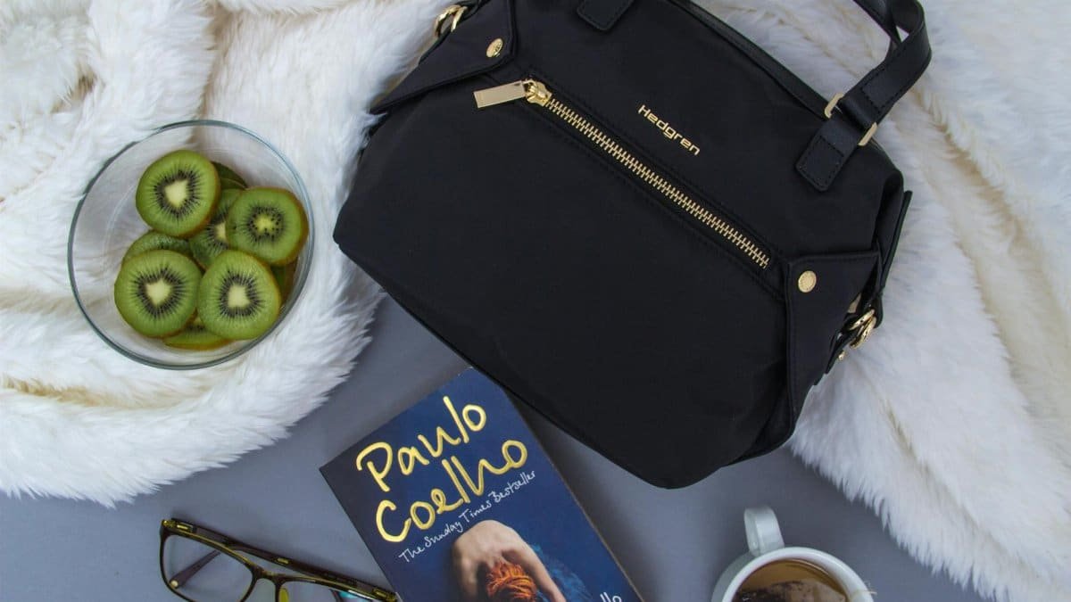 From above of classic bag and book with eyeglasses composed with bowl of fresh sliced kiwi and cup of tea and placed on gray bed with white fur blanket