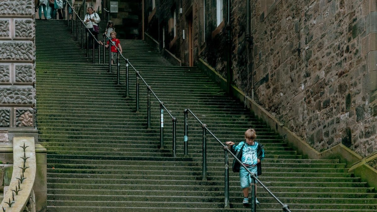 A child in a blue outfit walks down a moss-covered urban stairway, holding the handrails.
