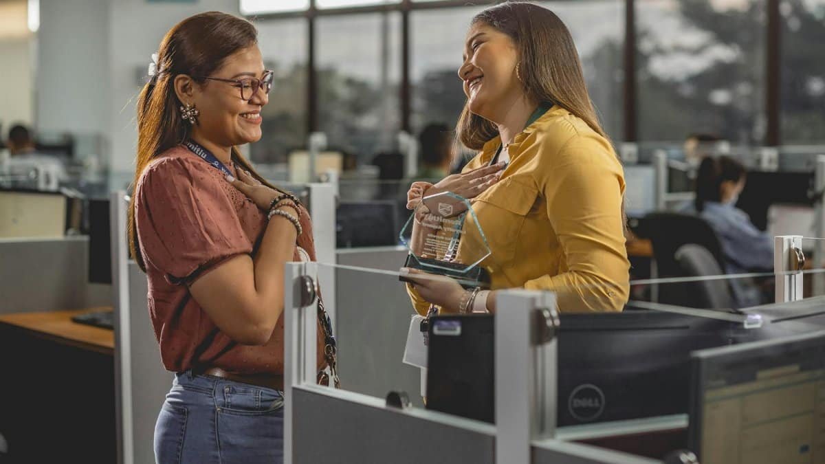 Two women smile and share a joyful moment receiving an award in a modern office setting.