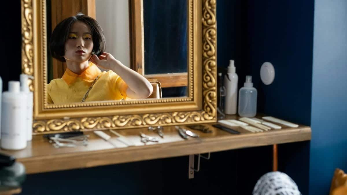 A woman in a yellow blouse elegantly gazing at her reflection in an ornate mirror.