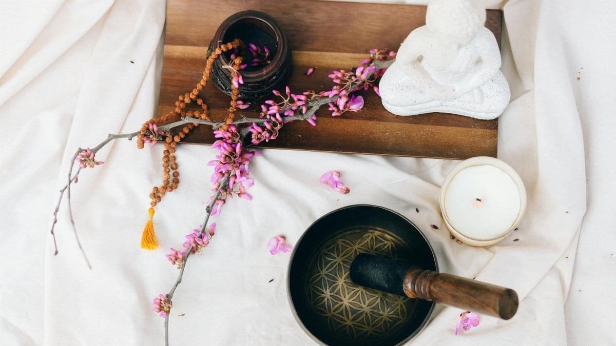Flat lay of a meditation setup with a singing bowl, candle, beaded mala, and flower branch.