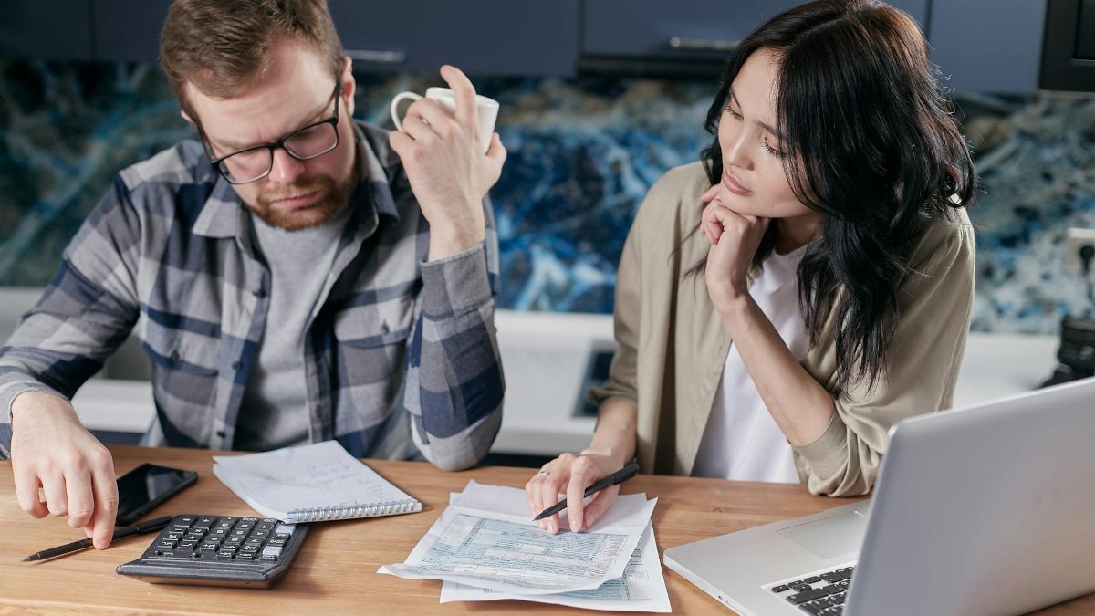 A couple reviews bills and documents on a laptop while discussing household finances.