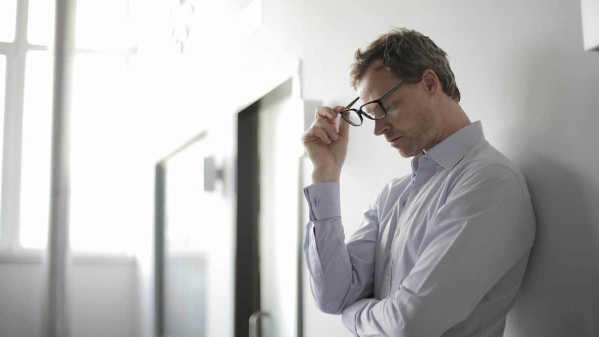 Thoughtful man in a bright room holding his glasses while leaning against a wall.