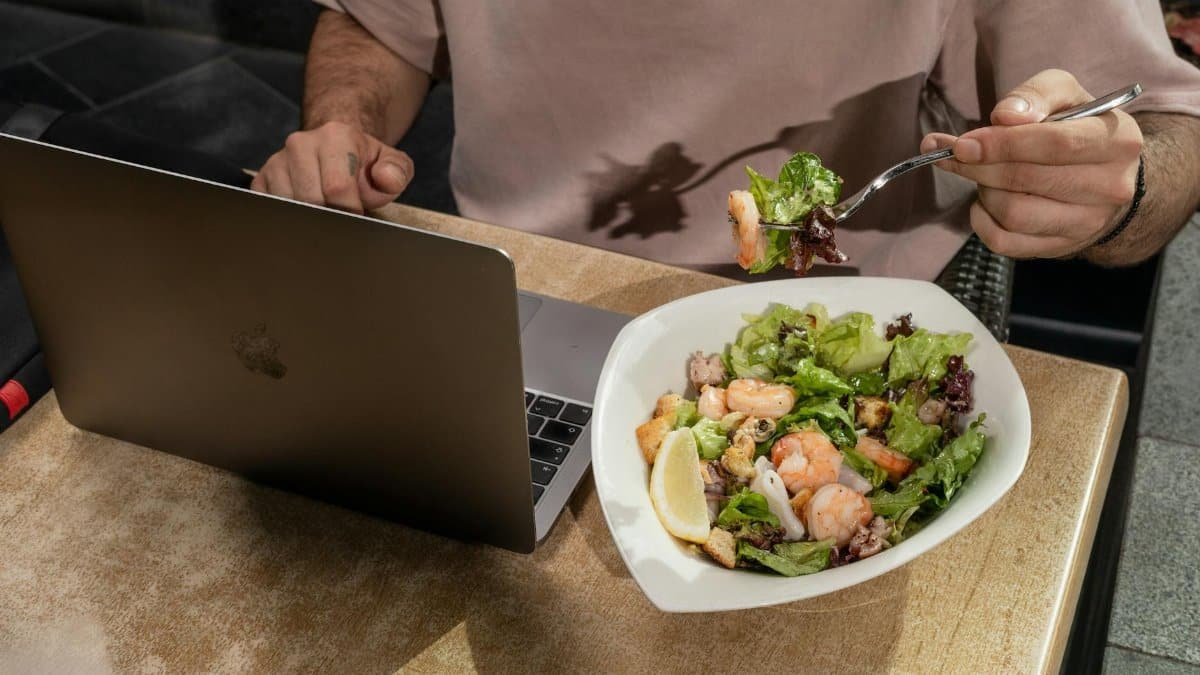 Adult man enjoying a shrimp salad while working on a laptop indoors.