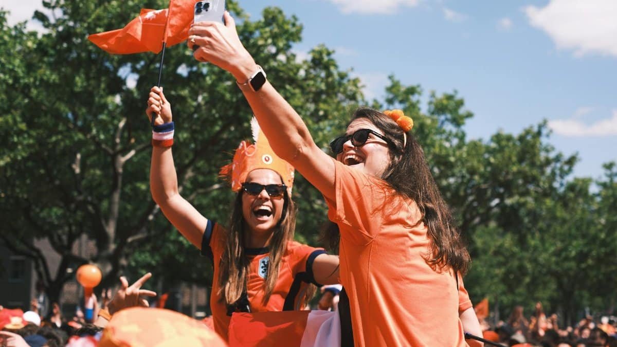 Excited Dutch football fans take selfies and celebrate outdoors wearing orange, waving flags.