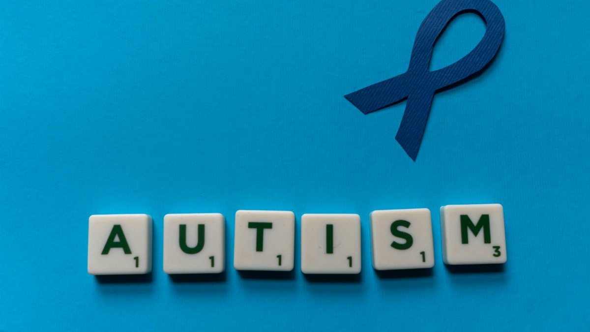 Flat lay of scrabble tiles spelling autism with a blue ribbon on a blue background.