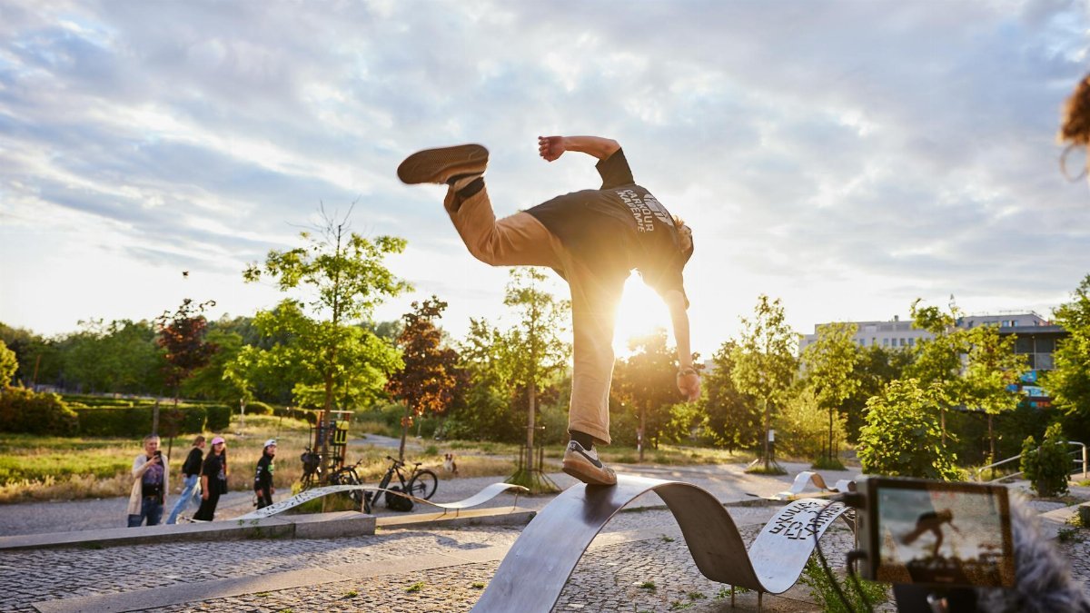 A teenager performs a skateboarding trick on a curved ramp in an urban park at sunset.