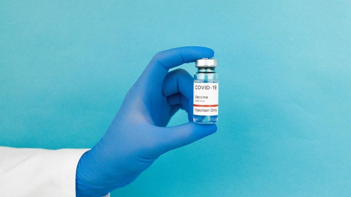 Close-up of a gloved hand holding a COVID-19 vaccine vial against blue background.