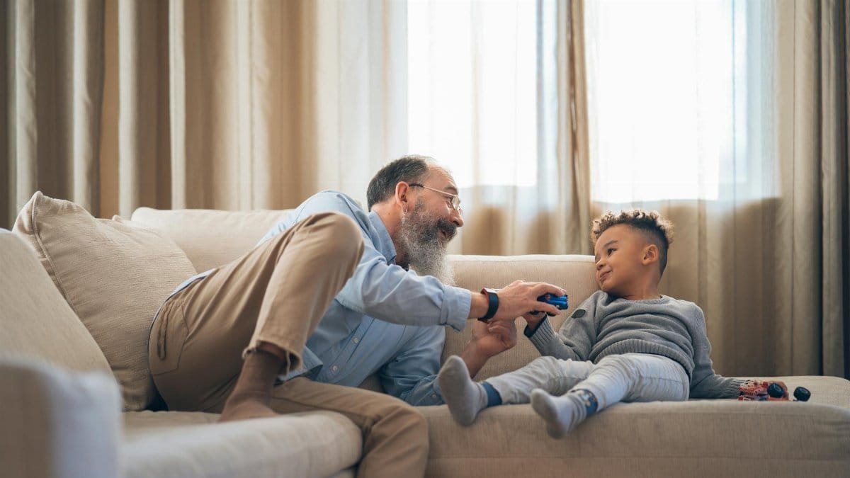 A heartwarming moment between grandfather and grandson laughing and playing on a cozy sofa indoors.