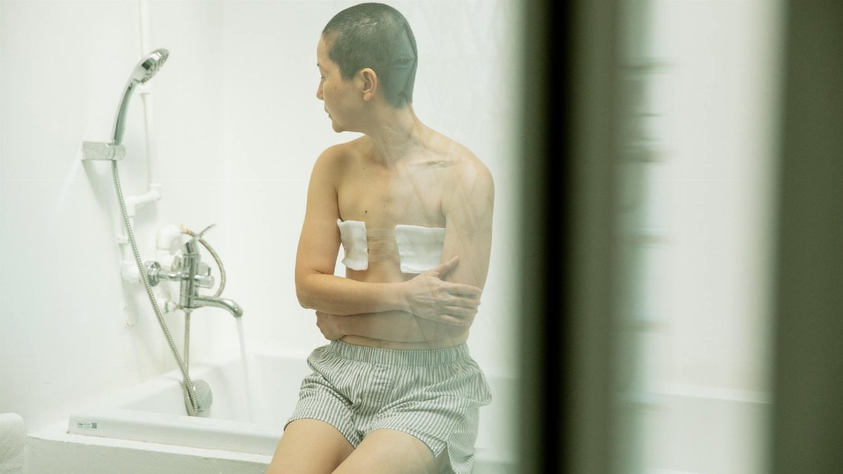Through glass pensive shirtless woman with short dark hair and bandages on breast after surgery sitting on bath in bathroom at home