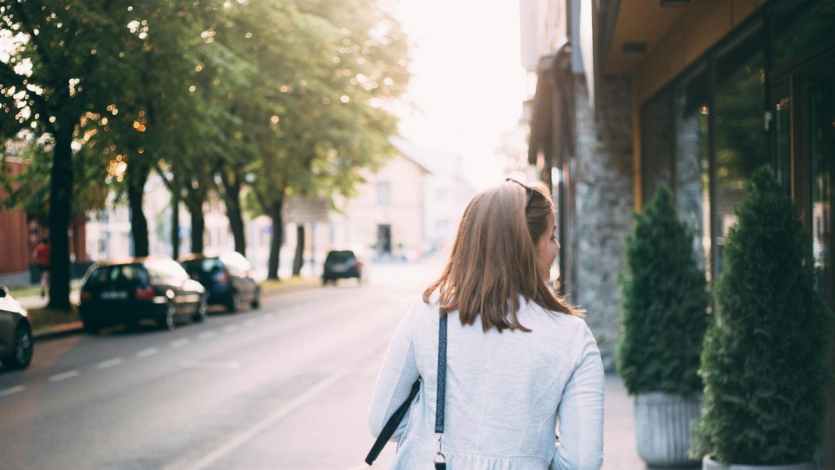 A woman walks down a quiet city street with trees and parked cars during daylight.