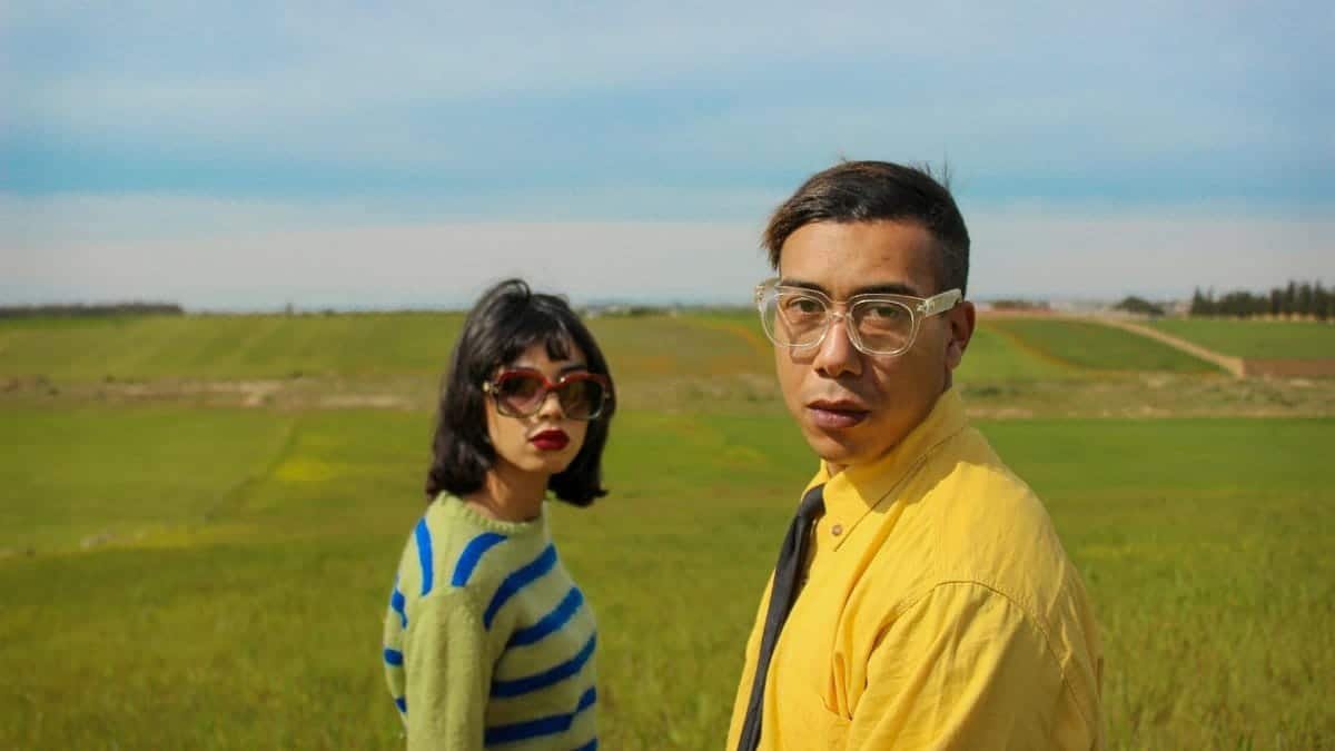 A duo in fashionable attire poses in a lush green field under a clear blue sky.