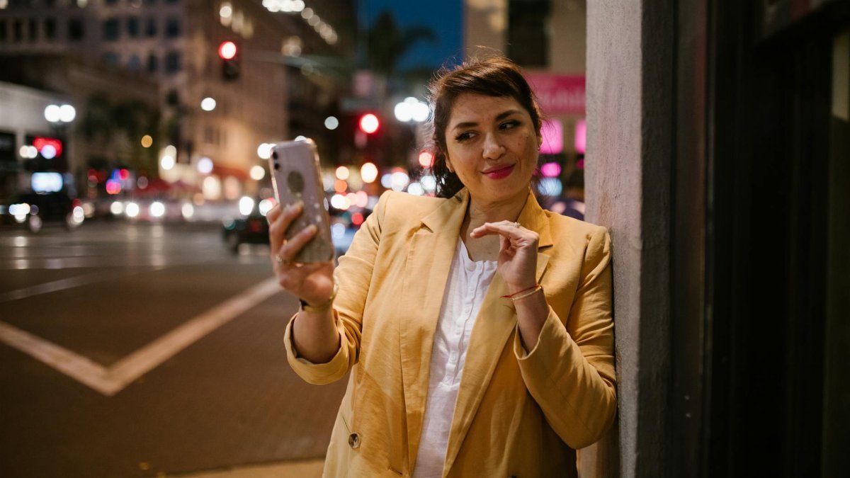 Woman in yellow blazer uses smartphone on a lively city street at night.
