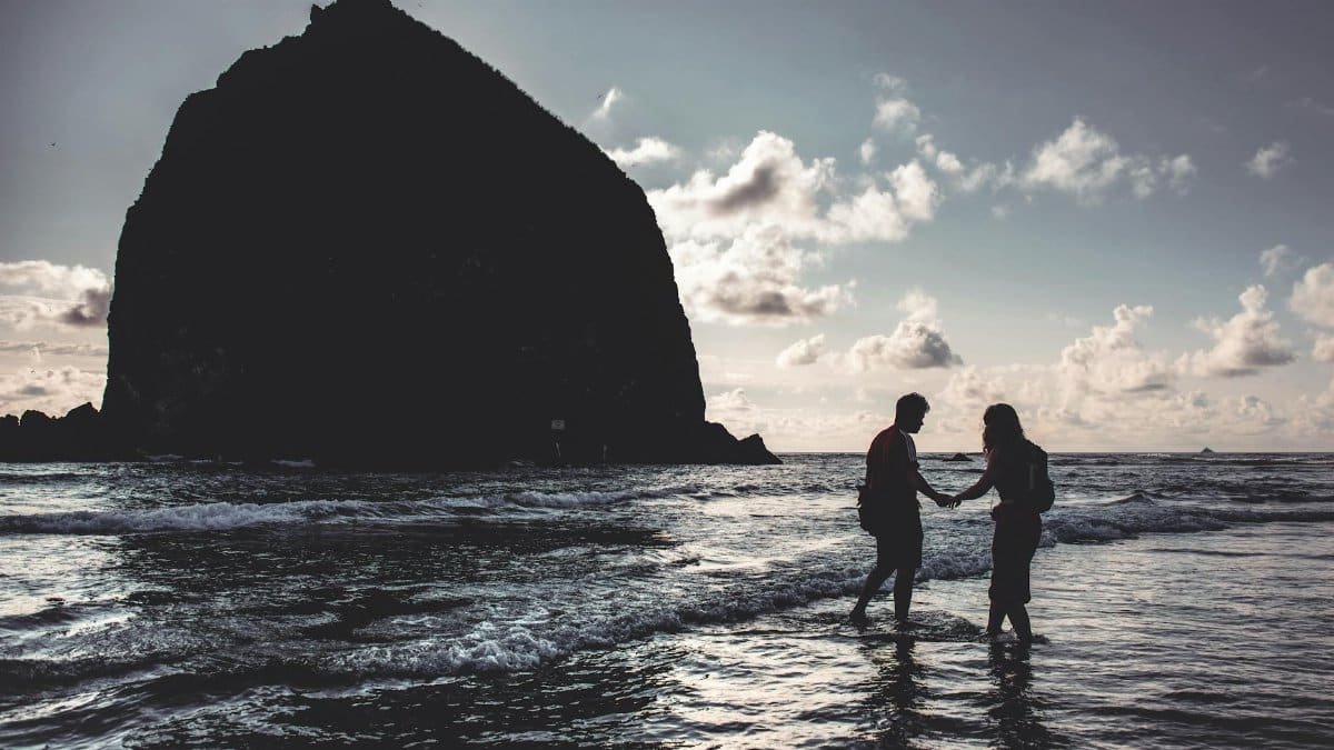 Silhouette of a couple holding hands at Cannon Beach during sunset, with Haystack Rock in view.