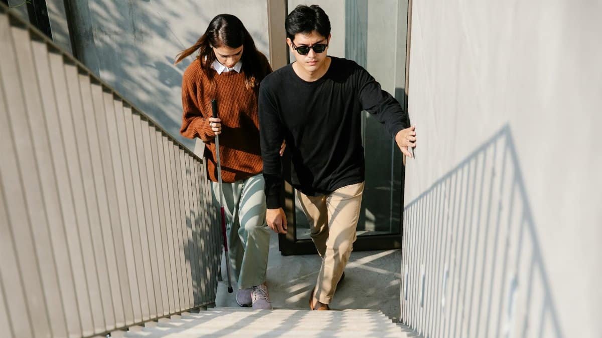 A man helps a visually impaired woman with a cane navigate stairs outdoors.