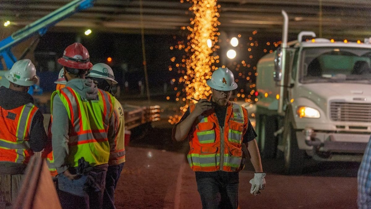 Construction workers in safety gear working at night with sparks flying under a bridge.