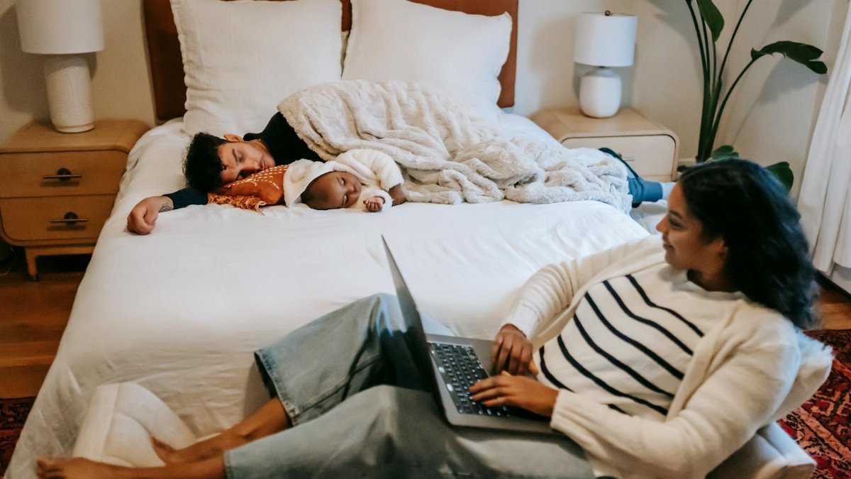 A mother works on a laptop while her family rests peacefully on a bed in a cozy bedroom.
