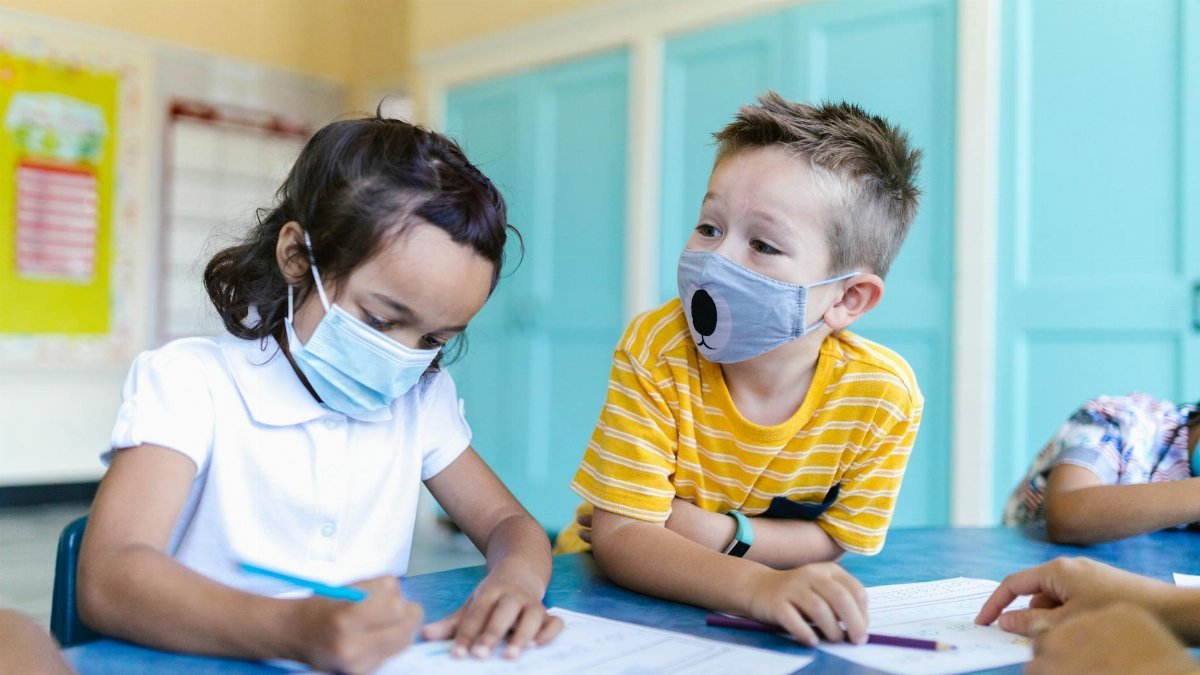 Two kids wearing masks focus on their work at a classroom table.