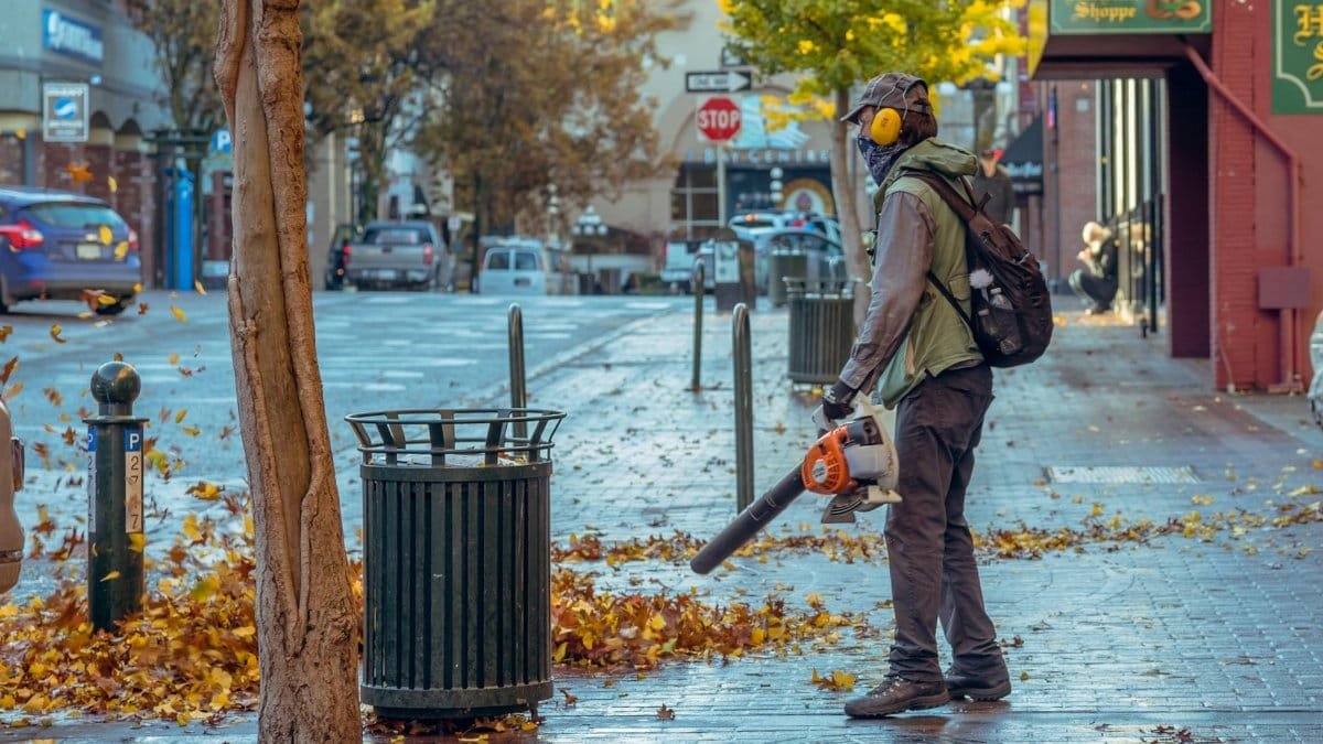 Street worker using a leaf blower during autumn in a city setting, surrounded by fallen leaves.