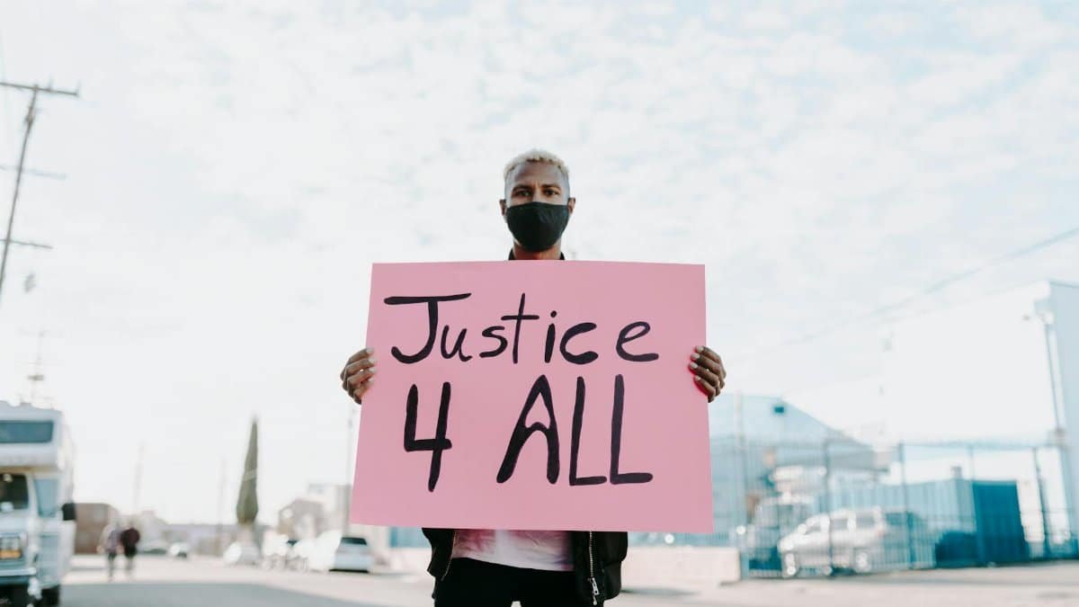 Man holding a 'Justice 4 All' sign while wearing a face mask during an outdoor protest.
