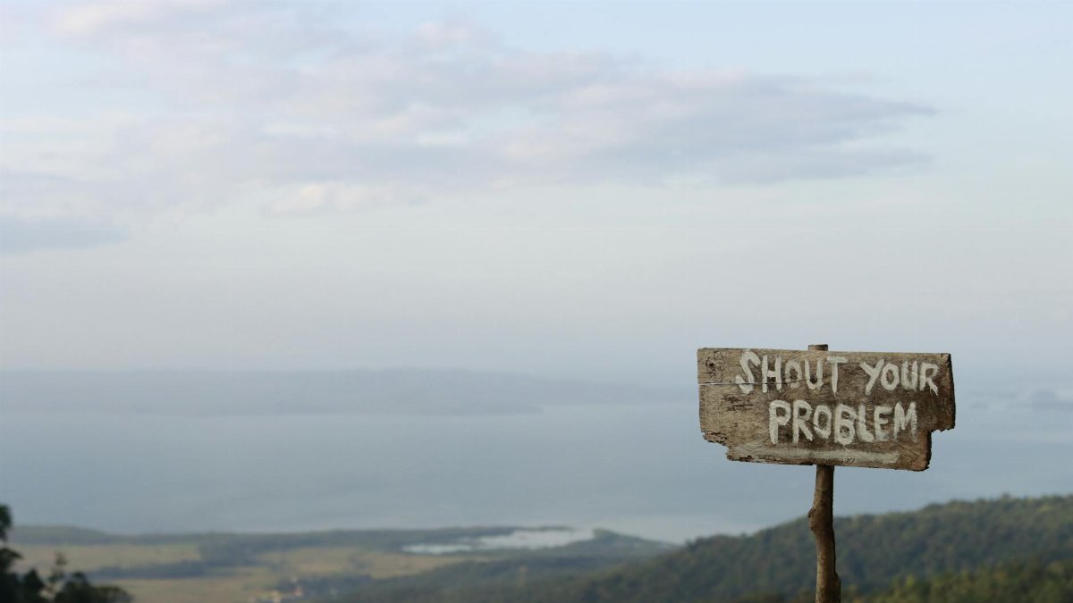 Wooden sign inscribed with 'Shout Your Problem' facing a beautiful mountain landscape under a blue sky.