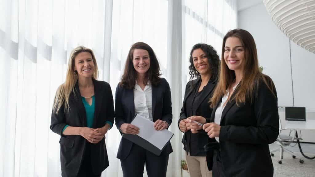 Four professional women in business attire smiling in a bright, modern office.