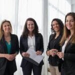 Four professional women in business attire smiling in a bright, modern office.