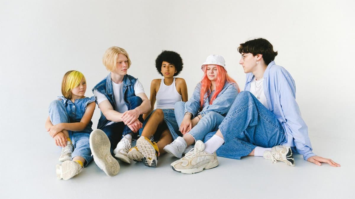 Full body group of thoughtful multiethnic teenager friends in jeans clothes sitting on floor and speaking with each other against white background