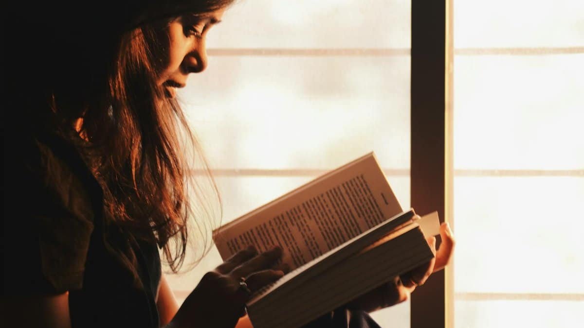 A woman engrossed in reading a book by a sunlit window creates a serene atmosphere.