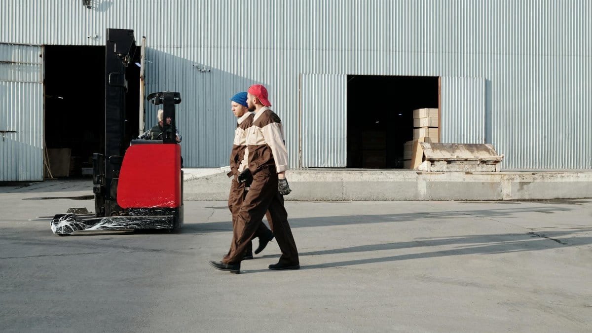 Two workers in uniforms walking outdoors near a forklift at a warehouse.