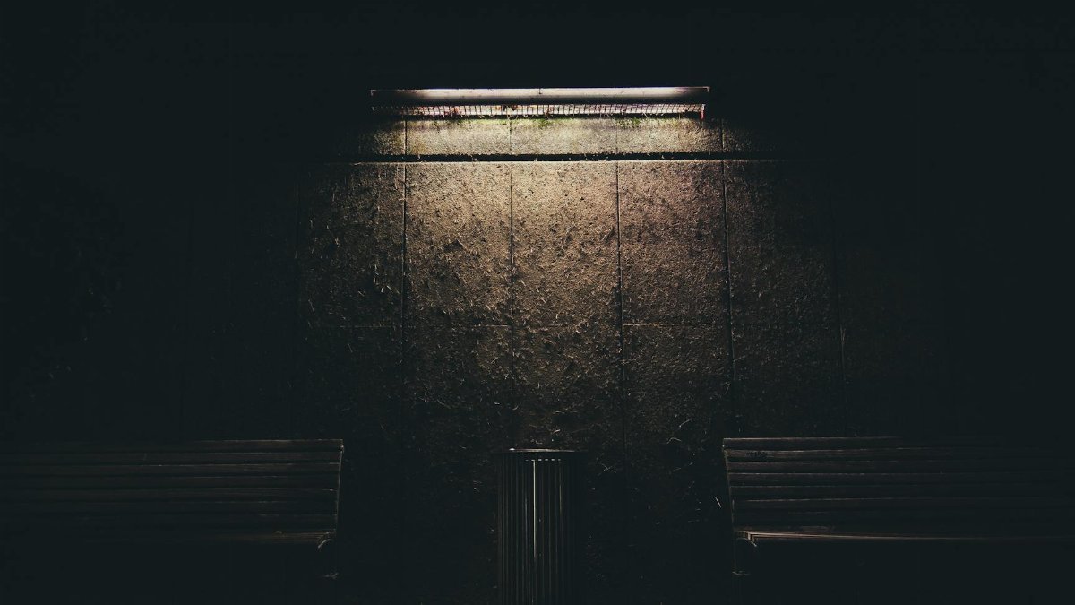 Dark urban night scene with benches and a fluorescent lamp casting soft light on the wall.