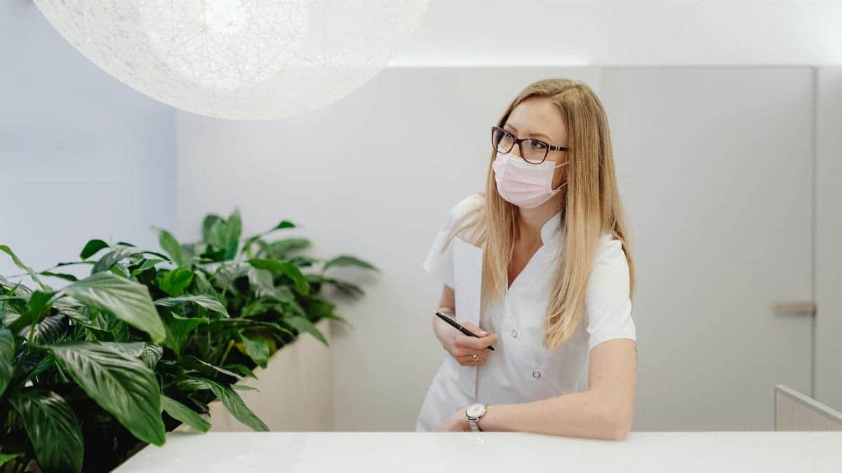 A female healthcare worker in a clinic wearing a face mask, standing at the reception desk.